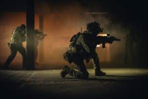 Four soldiers in tactical gear engage in a smoky nighttime military operation indoors.